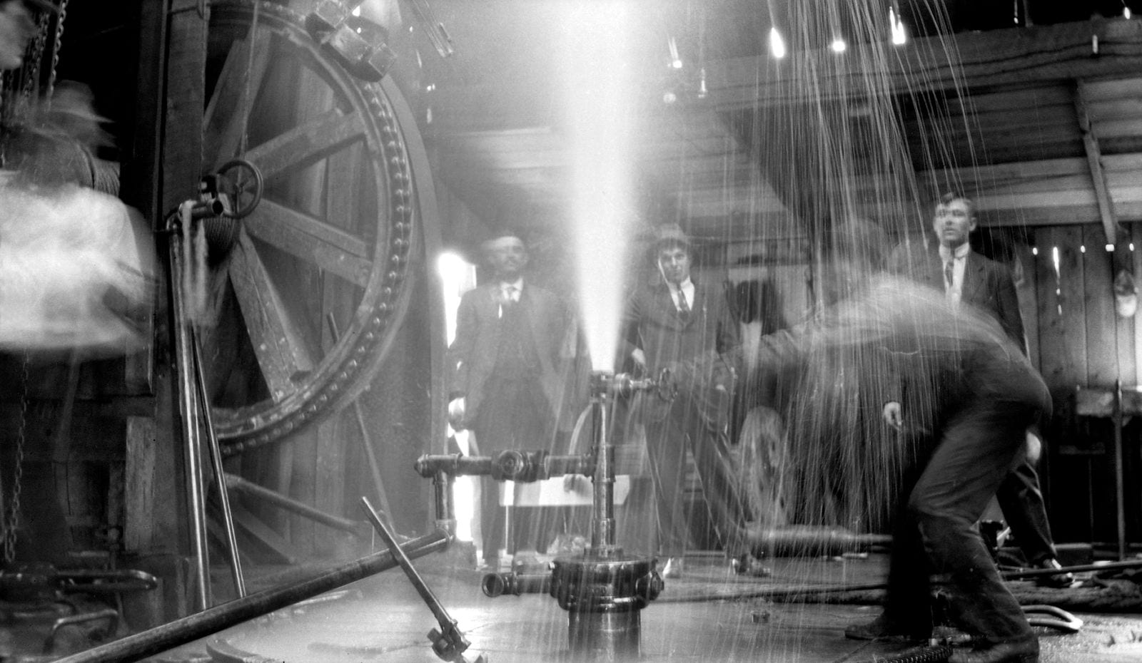 Dingman No. 1 oil well blowing inside its wooden derrick at Turner Valley, Alberta in 1914, with crew and onlookers in suits standing beside the wellhead